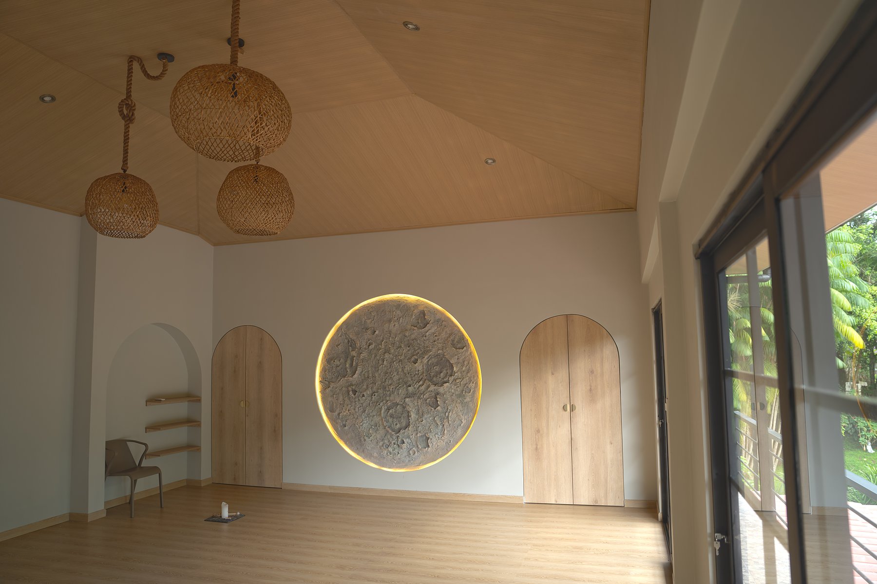 Wide corner view of the new practice room — the full vaulted ceiling, three rattan pendants, the lunar relief on the back wall, and floor-to-ceiling glass opening to the canopy on the right