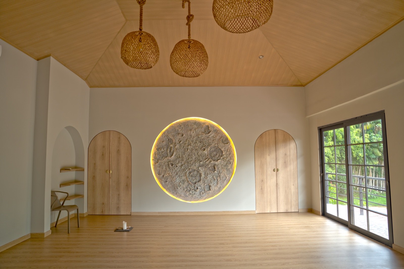 Head-on view of the new practice room — vaulted wood ceiling, the backlit lunar relief centered between two arched cedar doors, woven-rattan pendants overhead, and a reading chair with floating shelves on the left